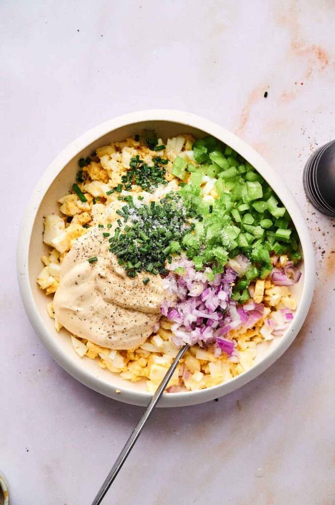 A bowl with chopped hard-boiled eggs, celery, red onion, chives, mayonnaise, and black pepper is ready to be mixed into a classic Egg Salad. A spoon rests in the bowl for easy mixing.