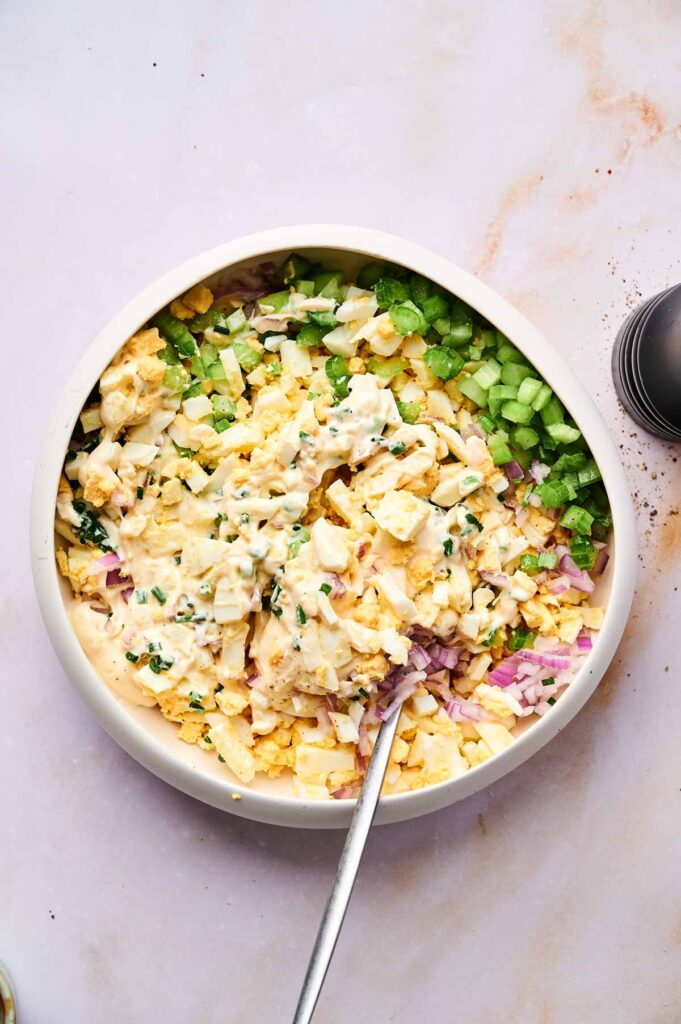 A bowl filled with classic egg salad—chopped hard-boiled eggs, celery, red onion, chives, and creamy mayonnaise—being mixed with a spoon.