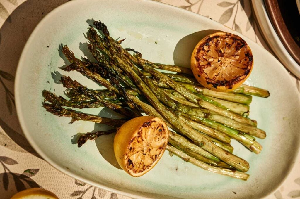 Grilled Asparagus spears served on a plate alongside two charred lemon halves for a smoky, citrusy finish.