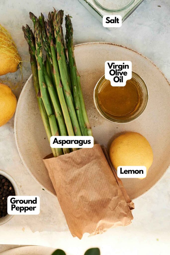 A bundle of grilled asparagus on a plate with a bowl of olive oil, a lemon, salt, and ground pepper displayed nearby.