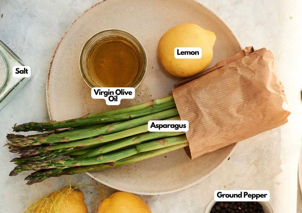 A plate with fresh asparagus, a lemon, a glass bowl of virgin olive oil, salt, and ground pepper, all labeled—perfect ingredients for making delicious grilled asparagus.