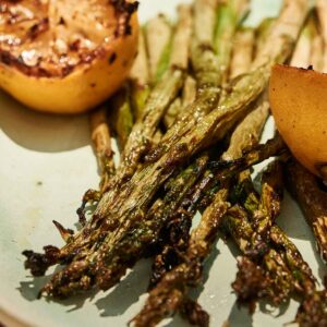 Grilled asparagus spears and two charred lemon halves are artfully arranged on a light-colored plate, highlighting the fresh flavors of perfectly grilled asparagus.