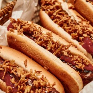 Three grilled hot dogs in buns topped with crispy fried onions and ketchup, arranged in a basket lined with parchment paper.