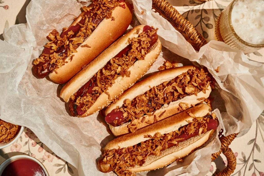 Three grilled hot dogs topped with crispy fried onions and ketchup are arranged in a basket lined with parchment paper, accompanied by a drink and condiments nearby on a floral tablecloth.