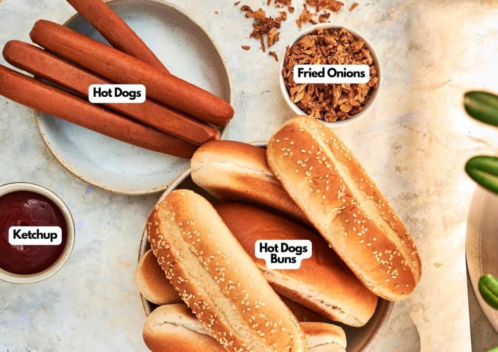 A plate of grilled hot dogs, a bowl of ketchup, a bowl of fried onions, and a plate of sesame hot dog buns are arranged on a light surface with labeled text.