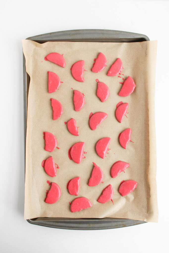 A baking tray lined with parchment paper holds rows of unbaked, pink, half-circle cookies arranged in a grid pattern, reminiscent of Ladybug Chocolate Covered Oreos awaiting decoration.