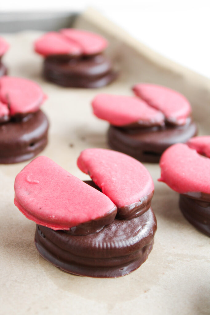 Ladybug Chocolate Covered Oreos, split in half and topped with pink heart-shaped decorations, are arranged on a parchment-lined baking sheet.