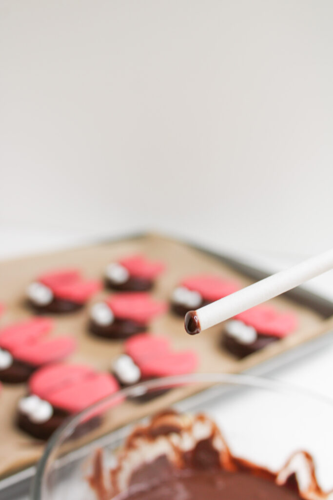 A close-up of a white lollipop stick dipped in chocolate, with heart-shaped, pink and brown cookies and Ladybug Chocolate Covered Oreos on a baking tray in the background.