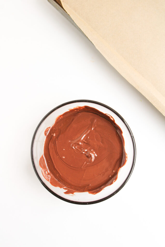 A glass bowl filled with melted chocolate is placed on a white surface next to a sheet of parchment paper, ready for making Ladybug Chocolate Covered Oreos.