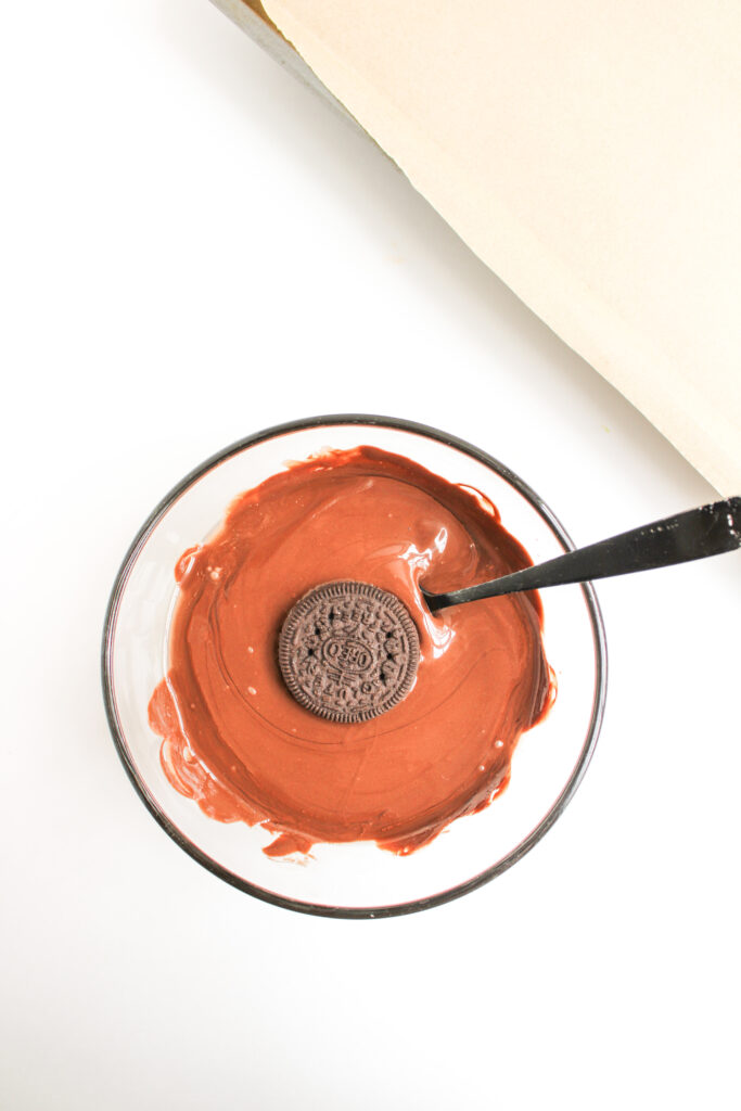 A Ladybug Chocolate Covered Oreo rests in a bowl of melted chocolate, with a spoon perched on the rim, ready for dipping.