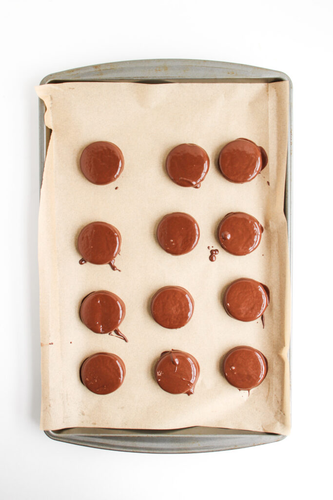 A baking tray lined with parchment paper holds twelve evenly spaced, round Ladybug Chocolate Covered Oreos, each dipped in chocolate and ready to enjoy.