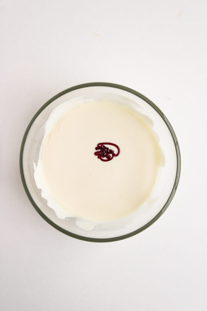 A glass bowl filled with white batter for Ladybug Chocolate Covered Oreos, with a small amount of red food coloring dropped in the center, on a white surface.