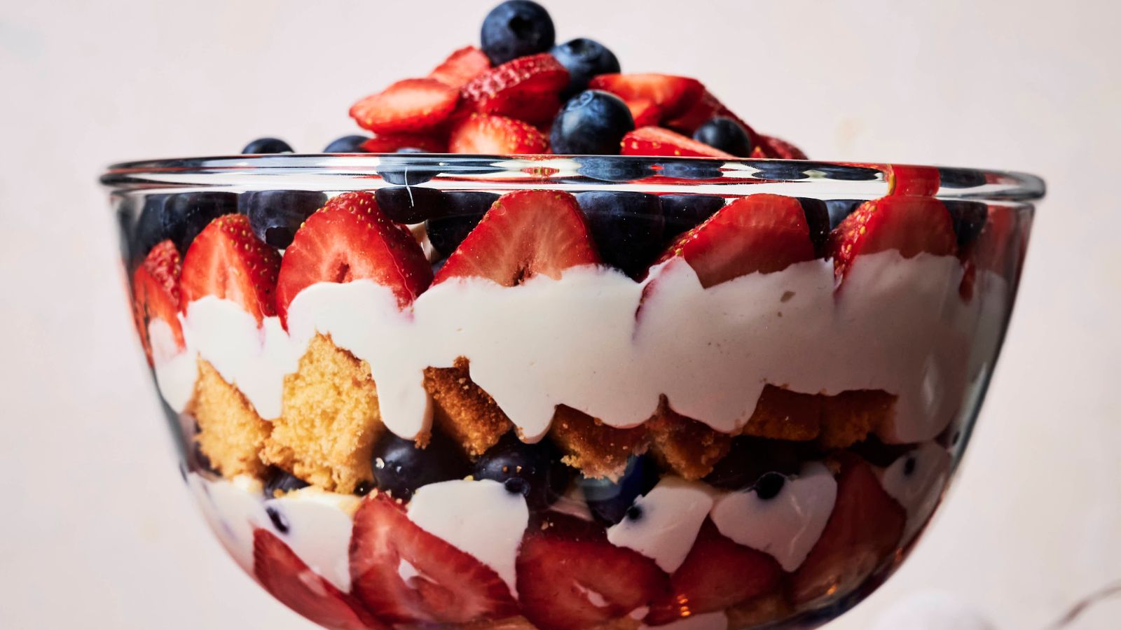 A glass bowl filled with layered dessert, featuring sliced strawberries, blueberries, whipped cream, and cake pieces.