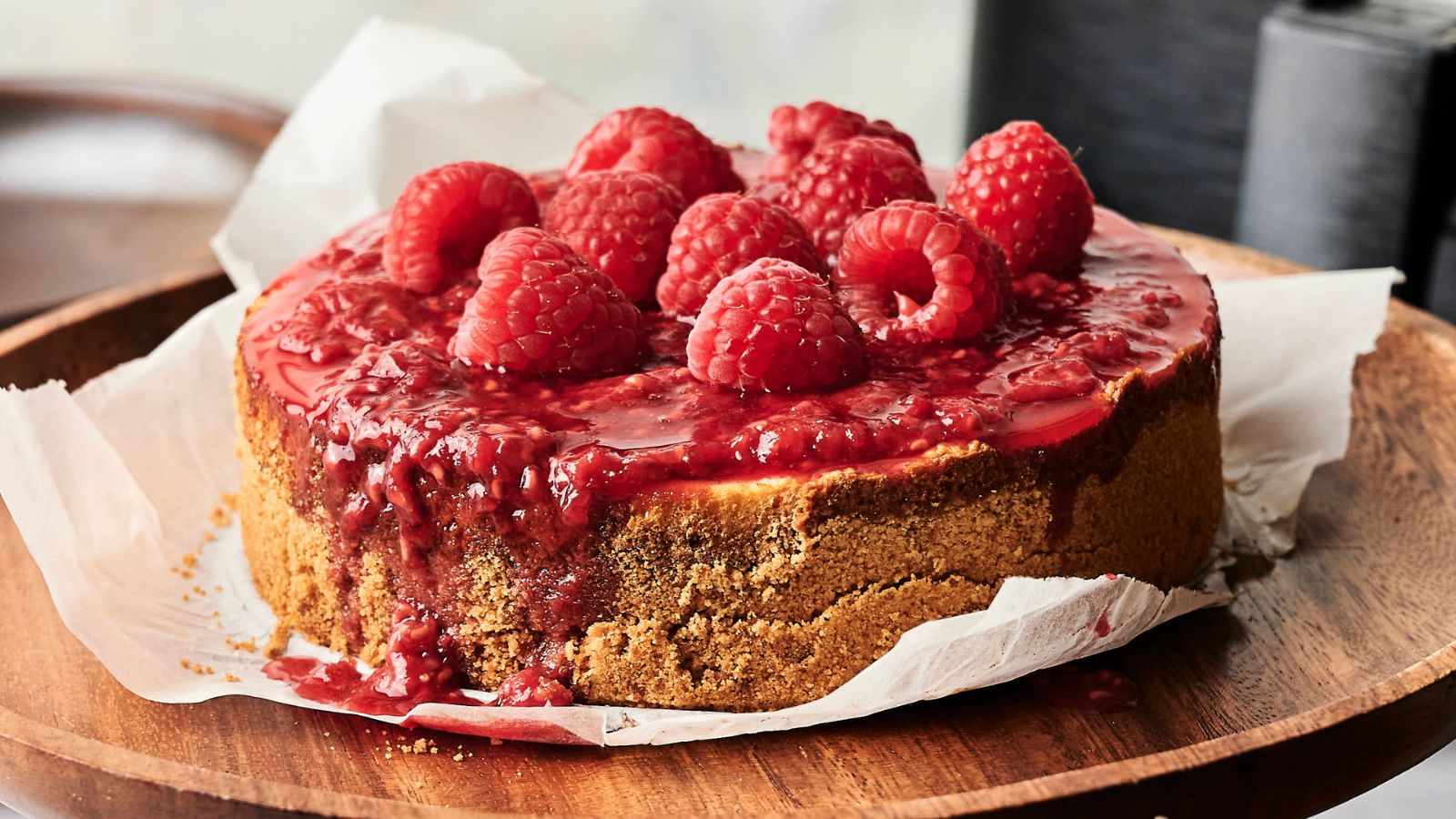 A cheesecake topped with raspberry sauce and fresh raspberries sits on parchment paper on a wooden plate.