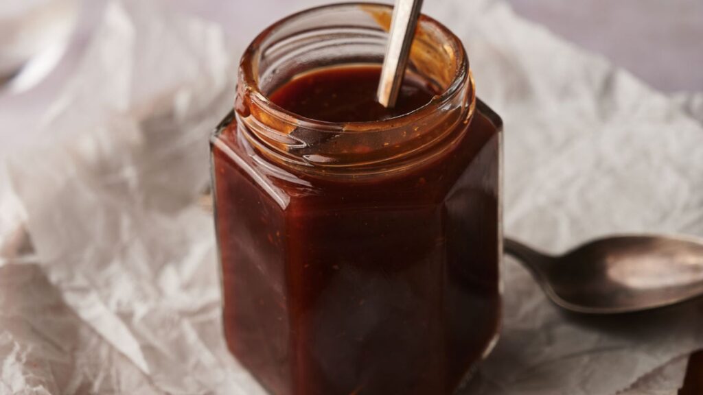 A hexagonal glass jar filled with dark brown sauce sits on crumpled parchment paper, with a metal spoon inside and another spoon nearby.
