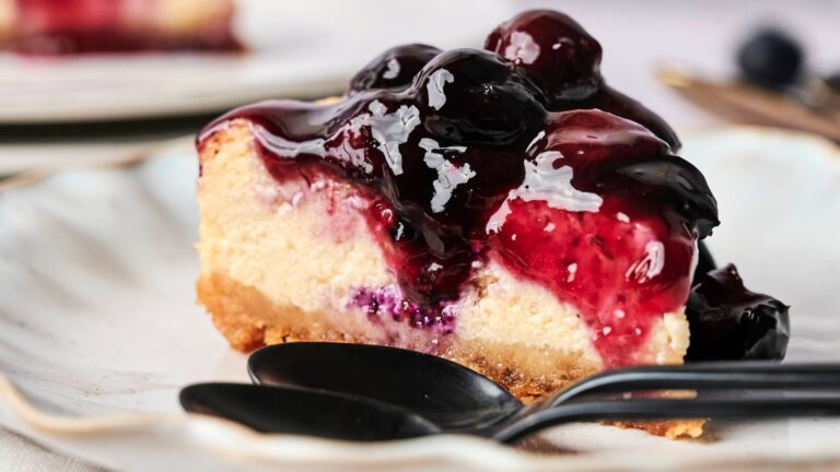 A slice of cheesecake topped with glossy blueberry sauce sits on a plate with two black spoons in the foreground.