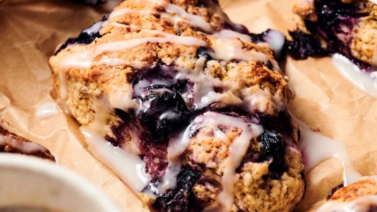 A close-up of a blueberry scone with white icing drizzled on top, resting on brown parchment paper.