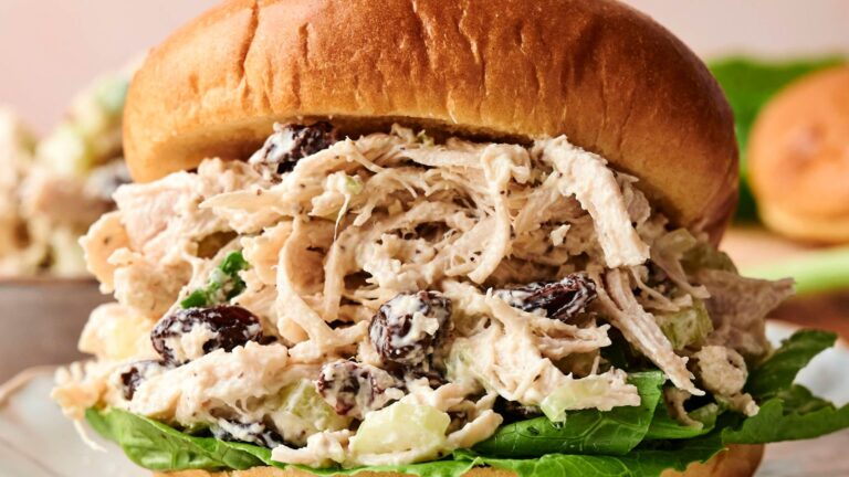 A close-up of a chicken salad sandwich with leafy lettuce, shredded chicken, celery, and raisins on a bun.