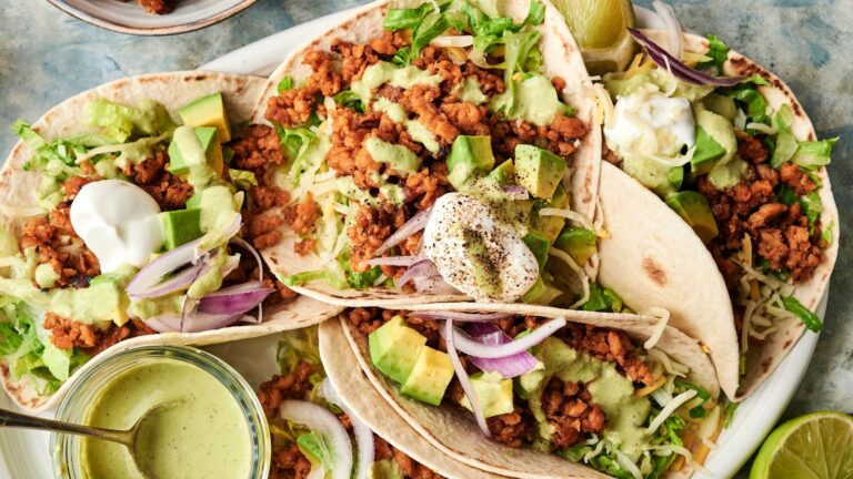 Three soft tacos filled with seasoned ground meat, lettuce, avocado, cheese, red onion, and topped with sour cream and green sauce, served on a white plate.