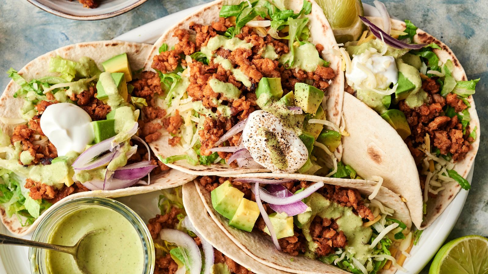 Three soft tacos filled with seasoned ground meat, shredded lettuce, diced avocado, red onion, shredded cheese, and topped with sour cream and green sauce sit on a plate.