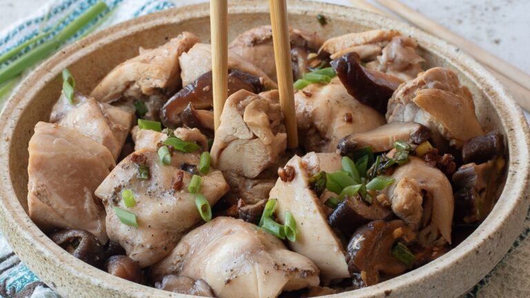 A bowl of cooked chicken pieces and mushrooms garnished with chopped green onions, with chopsticks picking up a piece.