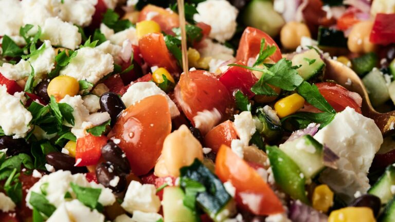 Close-up of a mixed cowboy caviar salad with tomatoes, chickpeas, black beans, corn, cucumber, red onion, feta cheese, and chopped parsley, being drizzled with dressing.