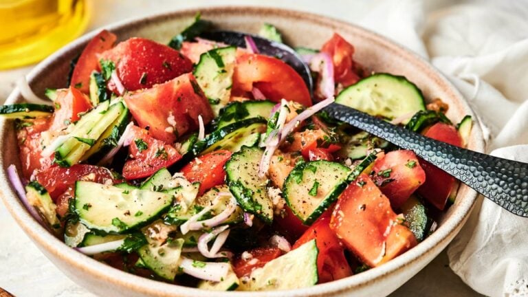 A bowl of tomato, cucumber, and red onion salad mixed with herbs and black pepper, with a serving spoon inside.