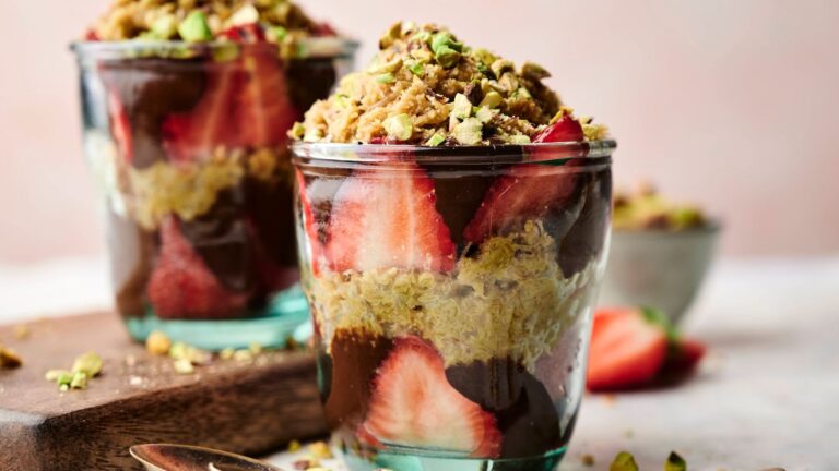 Two glass cups filled with layers of sliced strawberries, chocolate, crumbled pastry, and chopped pistachios on a light surface with a small bowl and spoon in the background.