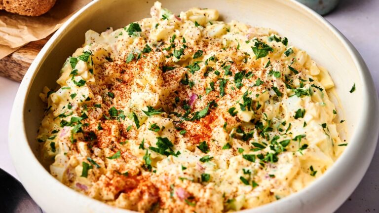 A bowl of creamy egg salad topped with chopped parsley and paprika, set on a light-colored surface.