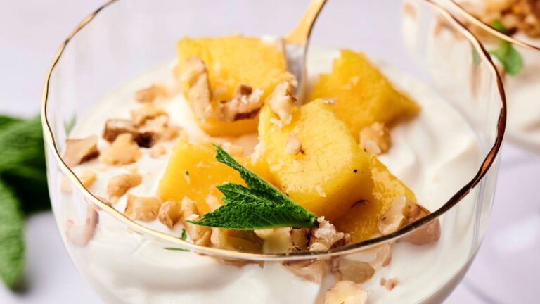 A glass cup filled with yogurt, topped with pineapple chunks, chopped nuts, and a sprig of fresh mint.
