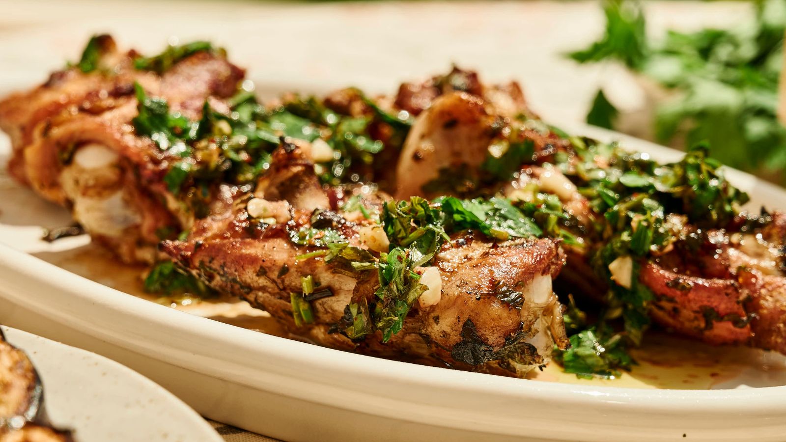 Grilled meat ribs garnished with chopped herbs and garlic, served on a white platter with fresh parsley in the background.