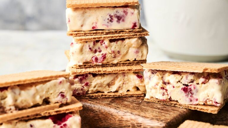 Ice cream sandwiches with pieces of fruit are stacked and arranged on a wooden board, with a blurred white background.