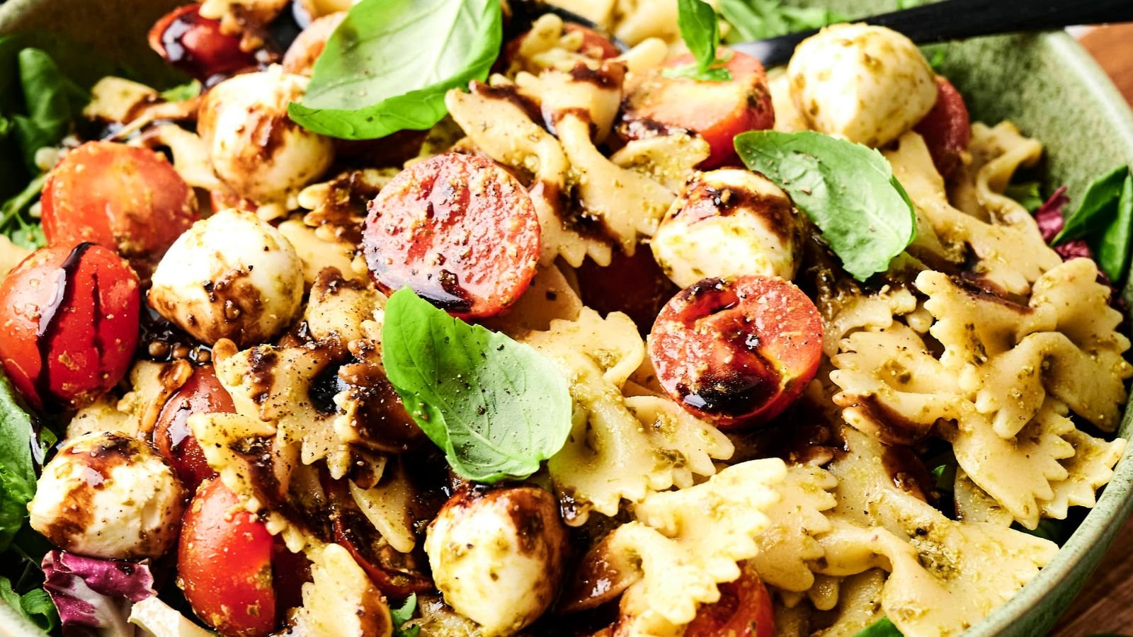 Bowtie pasta salad with cherry tomatoes, mozzarella balls, fresh basil leaves, and a drizzle of balsamic glaze in a green bowl.