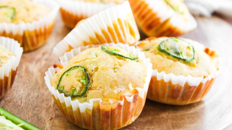 Two cornbread muffins topped with sliced jalapeños in white paper wrappers sit on a wooden surface, with more muffins in the background.