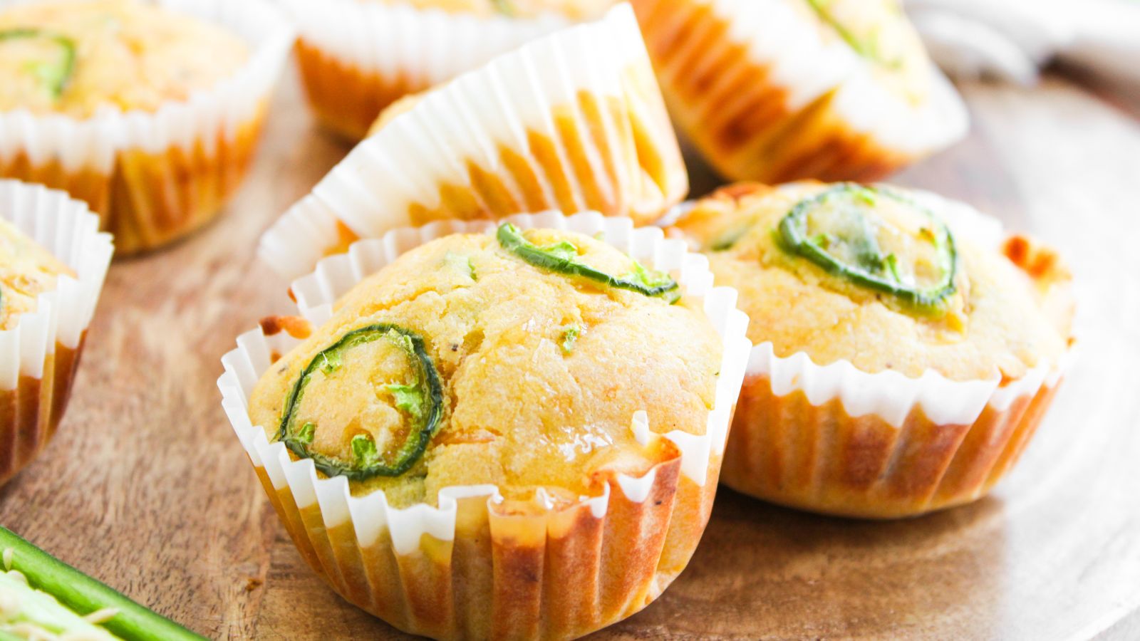 Cornbread muffins topped with sliced jalape&ntilde;os in white paper liners, arranged on a wooden surface.