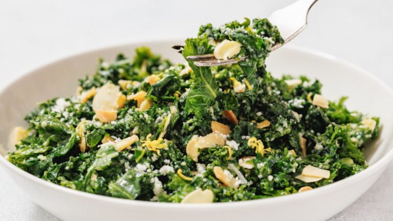 A bowl of kale salad with sliced almonds, grated cheese, and lemon zest; a fork lifts a bite from the bowl.