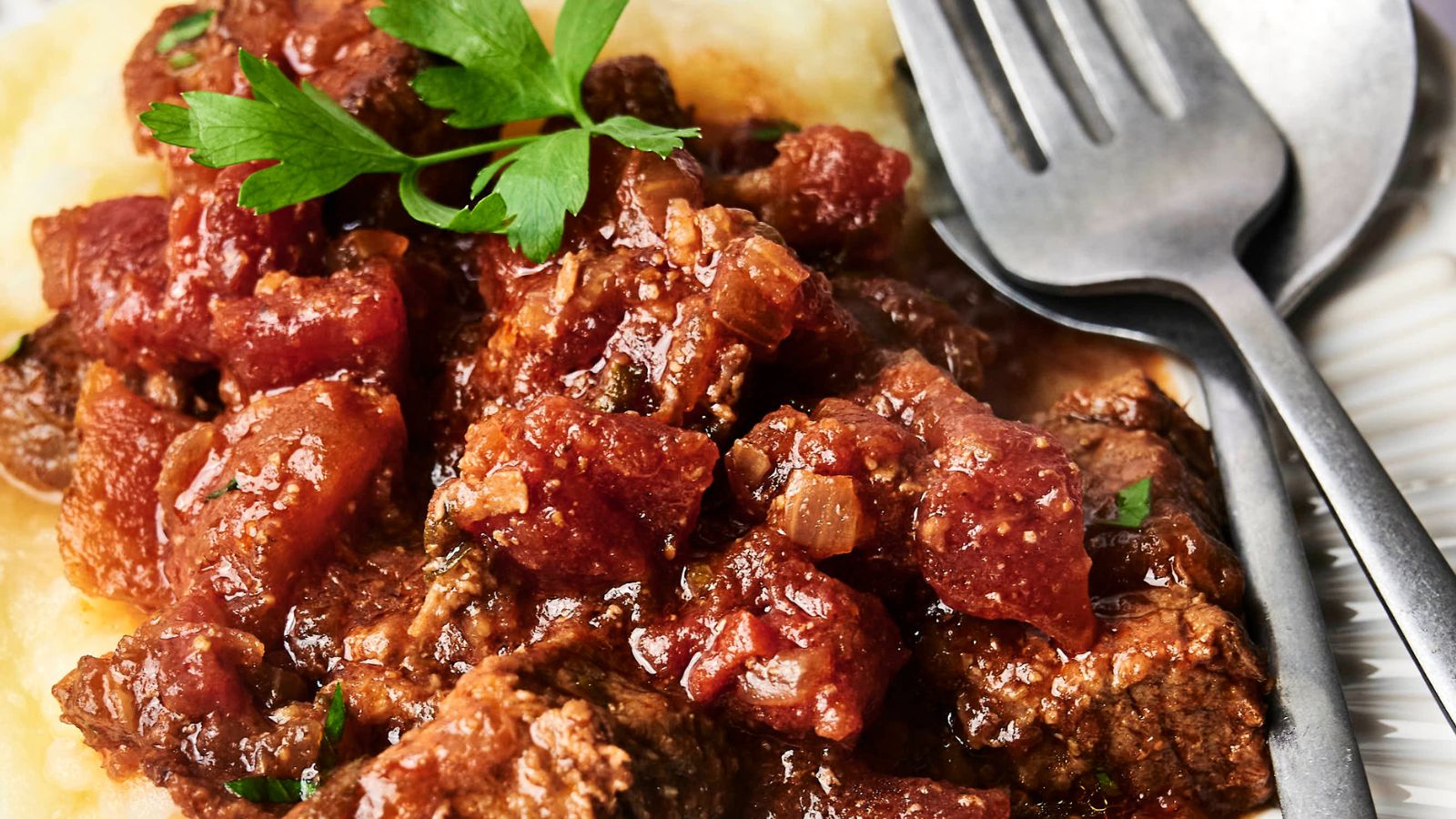 Close-up of beef stew with tomatoes and herbs served over mashed potatoes, garnished with parsley. A fork and spoon rest on the plate.