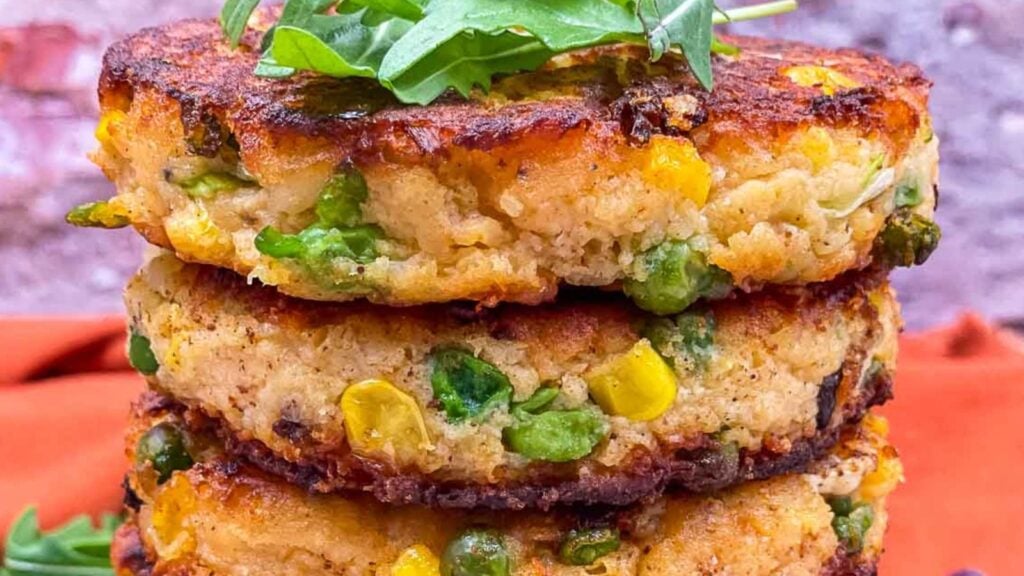 Three golden-brown vegetable patties with corn and peas are stacked on a plate, topped with arugula leaves.