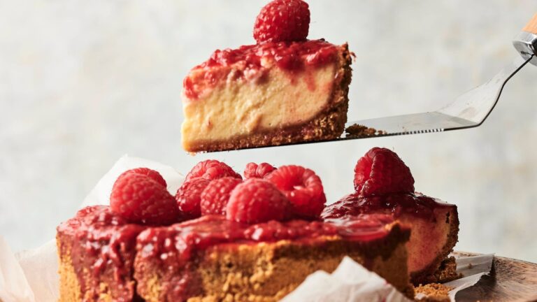A cheesecake topped with raspberry sauce and fresh raspberries, with one slice being lifted from the cake on a spatula.