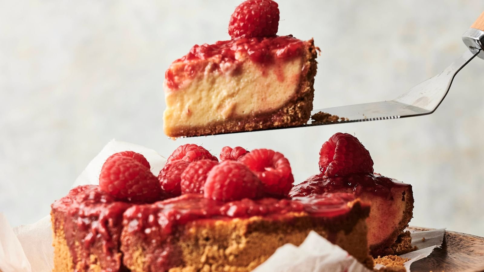 A slice of raspberry-topped cheesecake is being lifted from a whole cheesecake with a graham cracker crust.