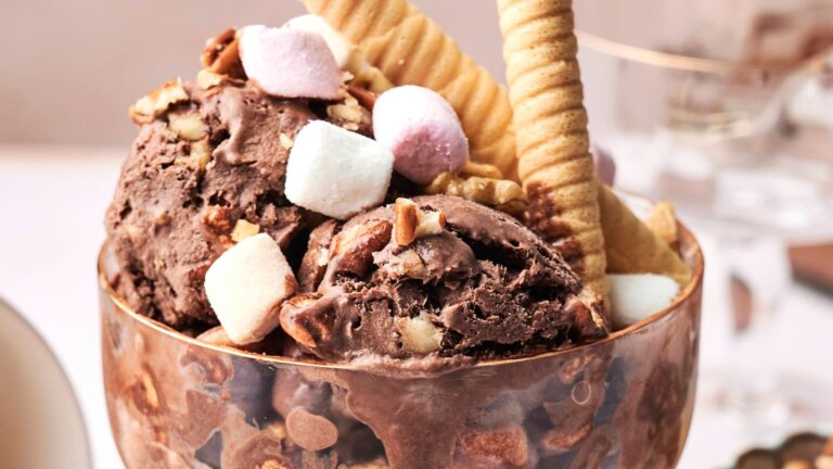 A glass bowl filled with scoops of chocolate ice cream, wafer rolls, waffle biscuits, chopped nuts, and marshmallows.