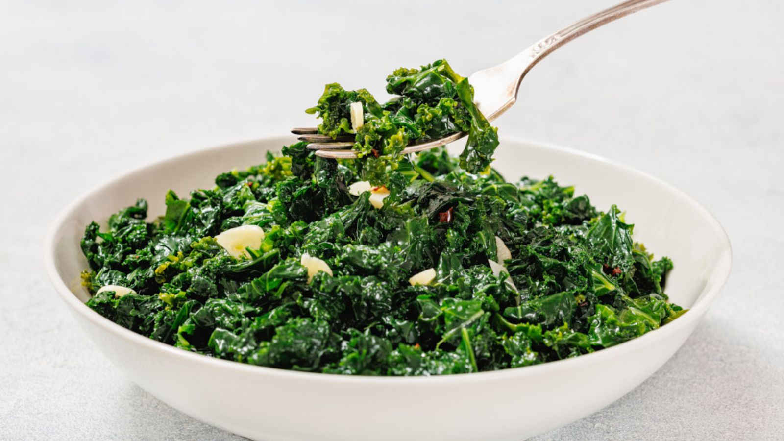 A white bowl filled with saut&eacute;ed kale and sliced garlic, with a fork lifting a portion from the bowl.