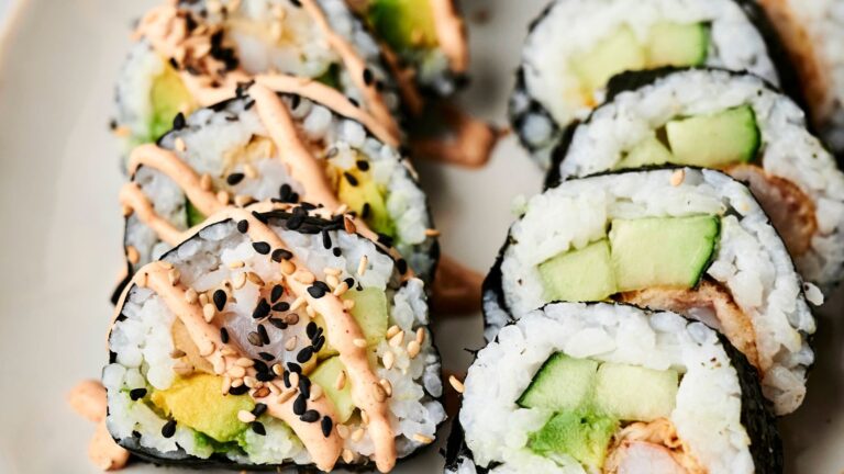 Close-up of sushi rolls with avocado, cucumber, and rice, topped with sesame seeds and a drizzle of spicy mayo on a beige plate.