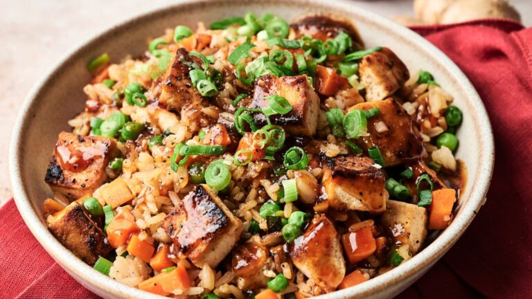 A bowl of fried rice with grilled tofu cubes, diced carrots, peas, chopped green onions, and a drizzle of sauce, served on a red cloth.