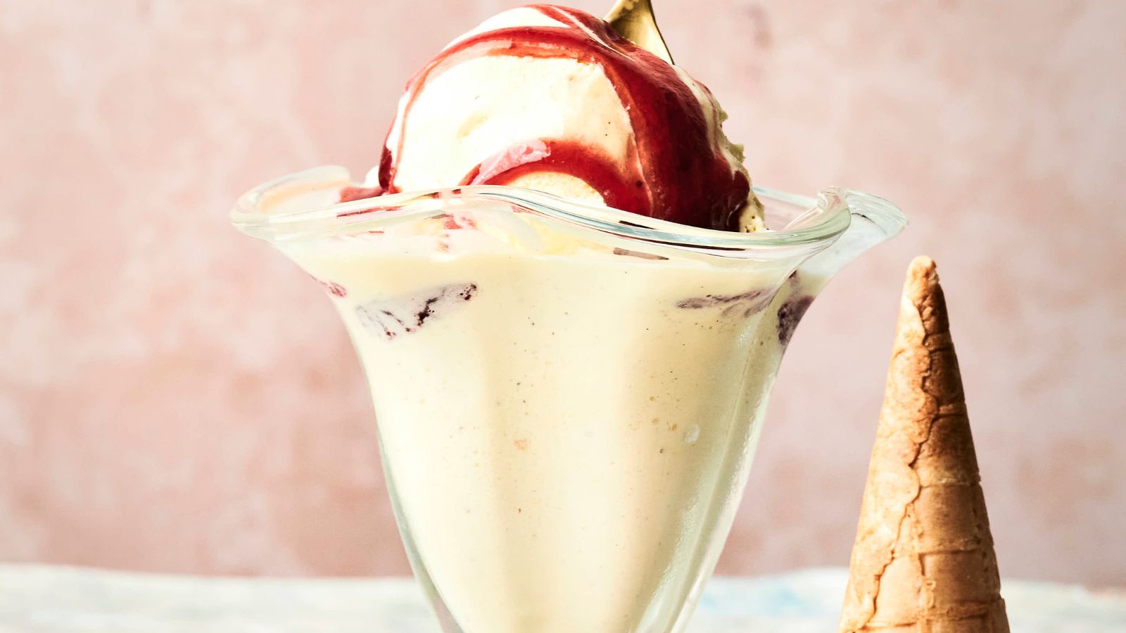 A glass dish with vanilla ice cream topped with red syrup, next to an empty ice cream cone, against a light pink background.