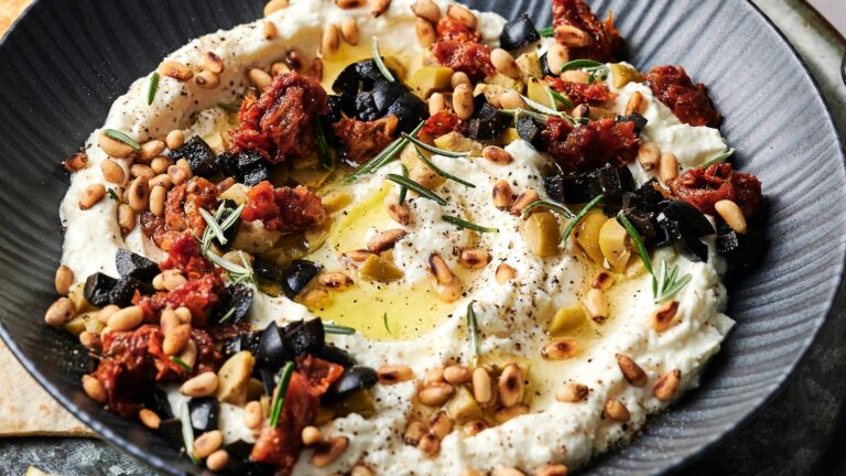 A bowl of whipped ricotta topped with pine nuts, chopped olives, sun-dried tomatoes, rosemary, cracked pepper, and a drizzle of olive oil.