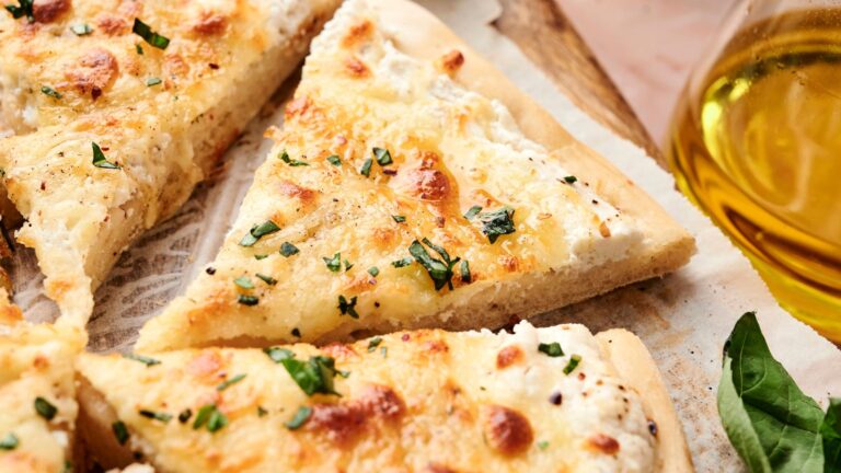 Close-up of sliced white pizza with melted cheese, herbs, and a thin crust on parchment paper, next to a glass container of olive oil and a basil leaf.