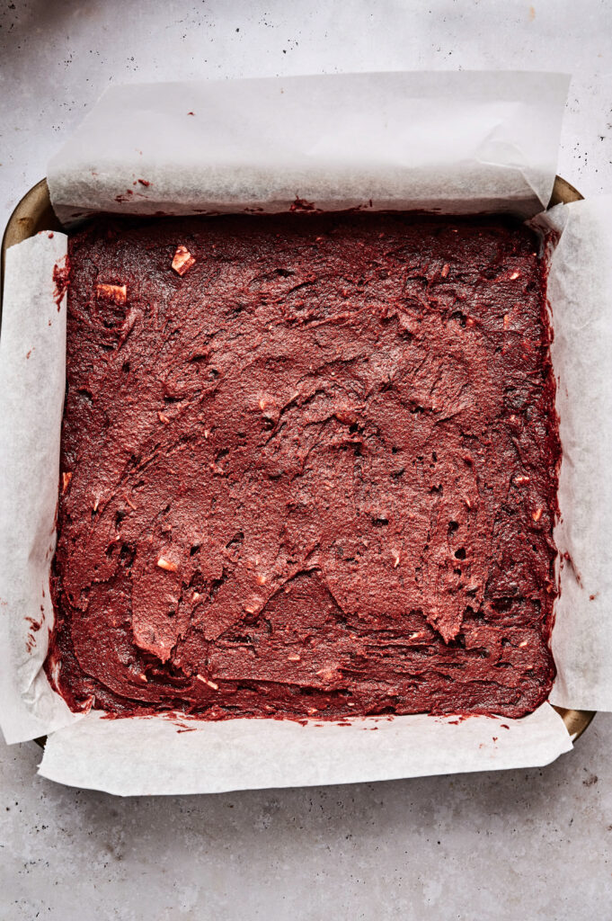 A square baking pan lined with parchment paper, filled with unbaked red velvet brownies batter.