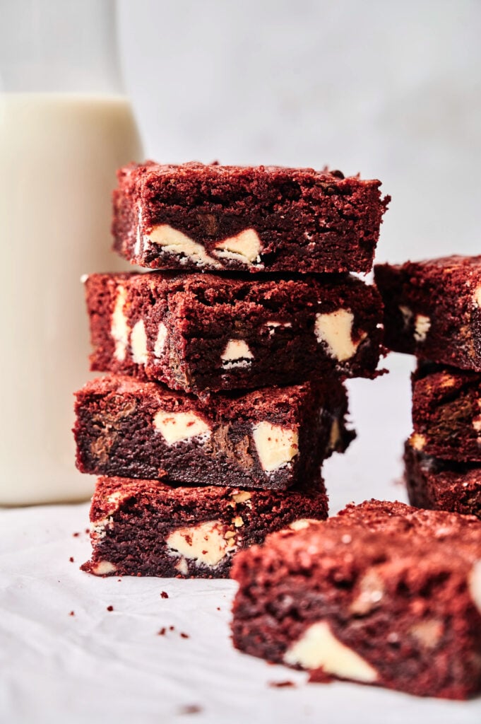 A stack of rich red velvet brownies with white chocolate chunks sits on a white surface next to a glass bottle of milk.