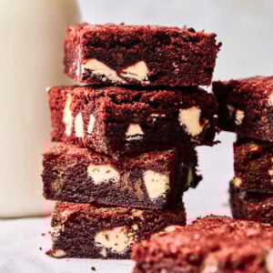 A stack of rich red velvet brownies with white chocolate chunks sits on a white surface next to a glass bottle of milk.
